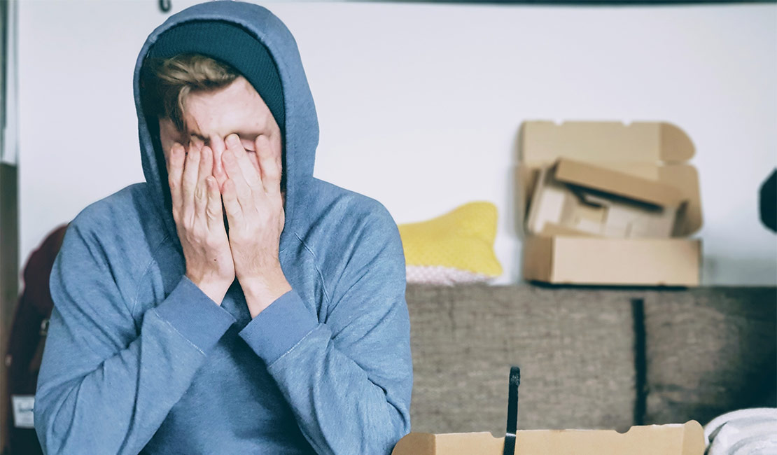 Man sitting on couch. He is rubbing his hands over his face in sleep deprivation. In the background there are boxes from moving.