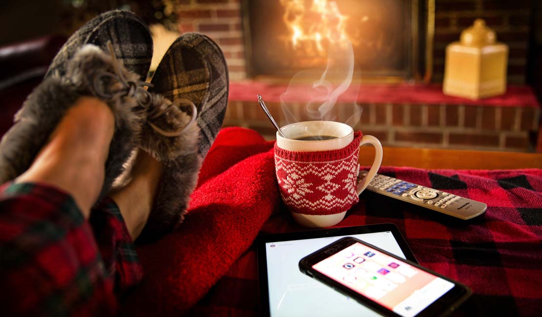 Warm cosy scene with lots of red and brown tones. Person wearing slippers on a stool next to a warm drink. Fireplace in the background.