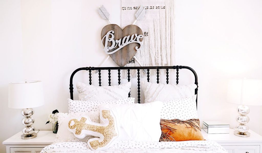 metal bed frame with white bedding and some throw pillows. Macrame and a sign that reads "brave" is against the wall above the bed.
