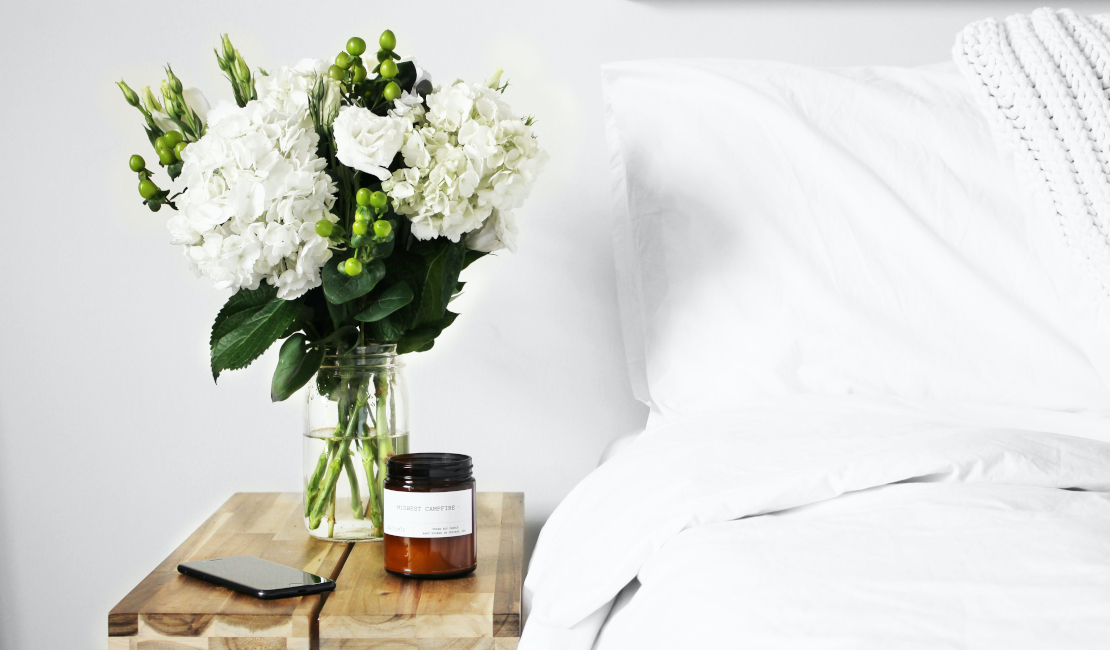 Made bed with white bedding and a wooden bedside pedestal with a vase of white flowers and a pot of face cream on the pedestal.