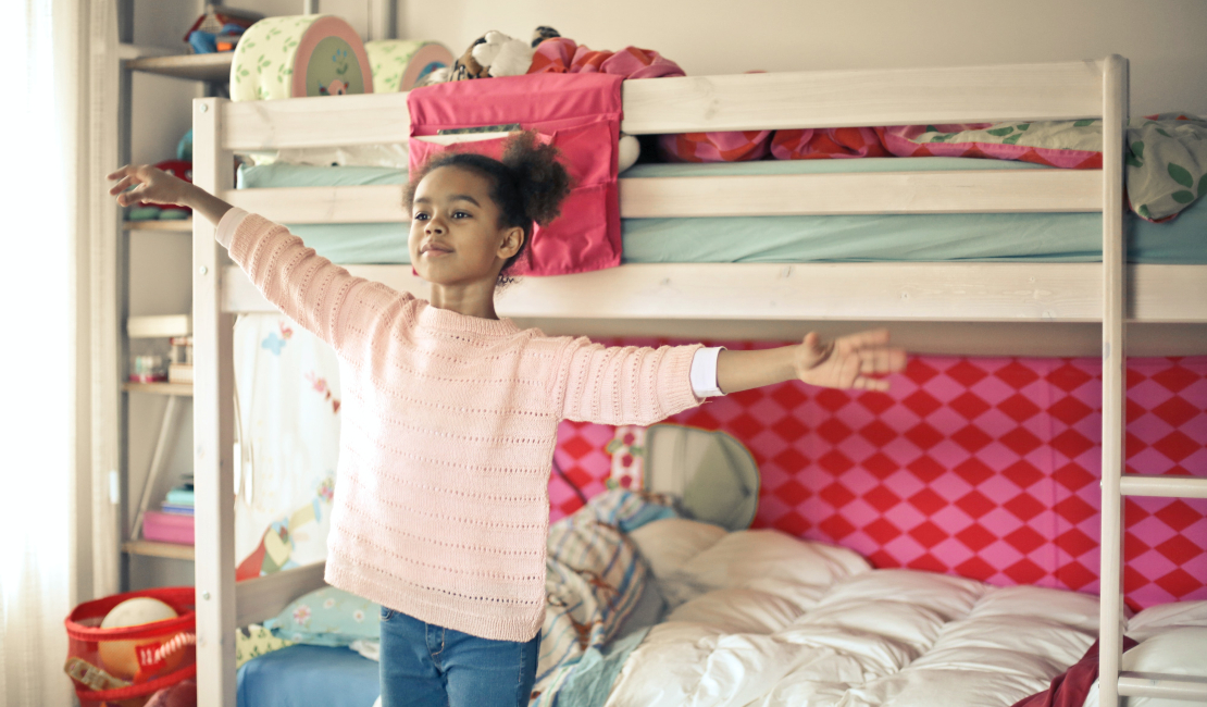 Bunk beds add flair to children's bedrooms. In this picture a girl is standing with outspread arms in front of her pink themed bunk bed. 