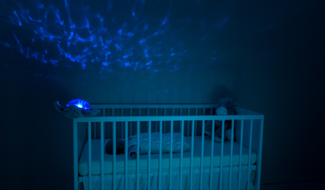 A baby cot in a dark room. There is a baby sleeping in the cot and a blue light casting starry patterns on the dark wall.