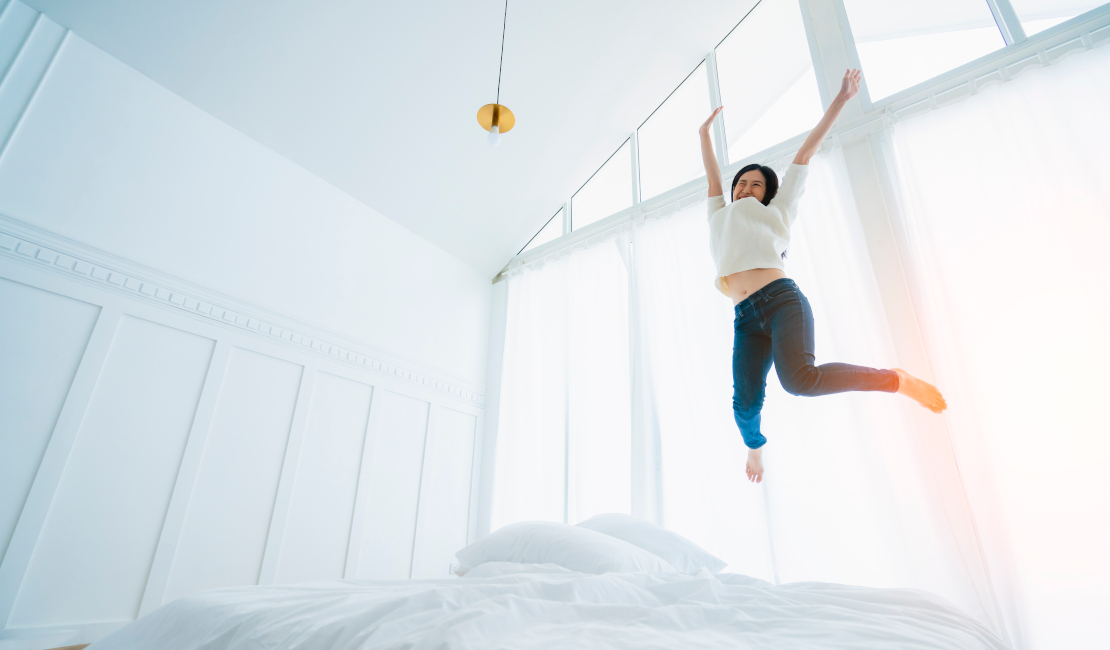 Woman jumping on a white bed in a white room with large windows.